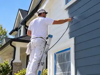 Professional painter spraying blue-gray exterior siding from a ladder